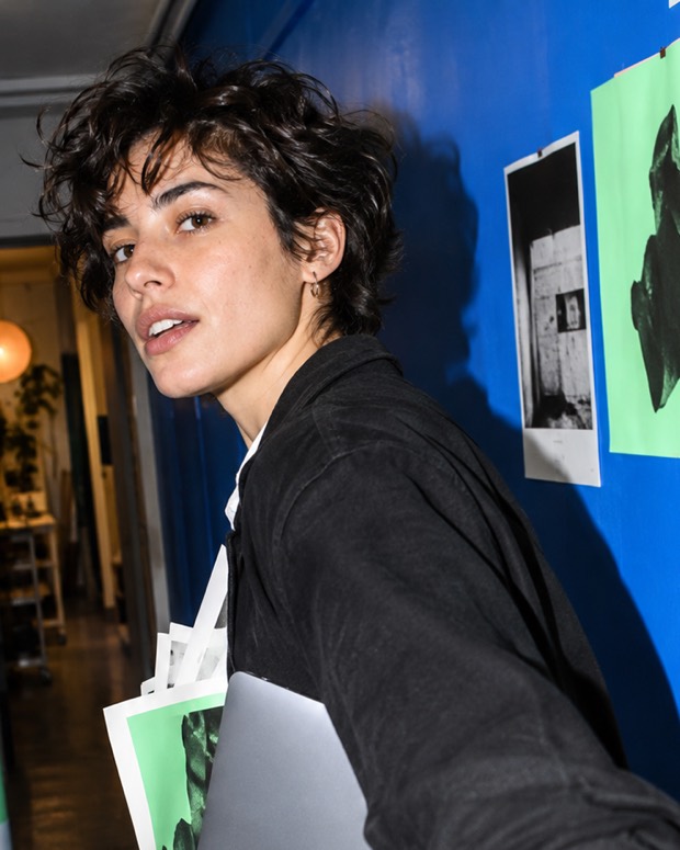 Someone crossing the studio with a laptop and a stack of prints under one arm