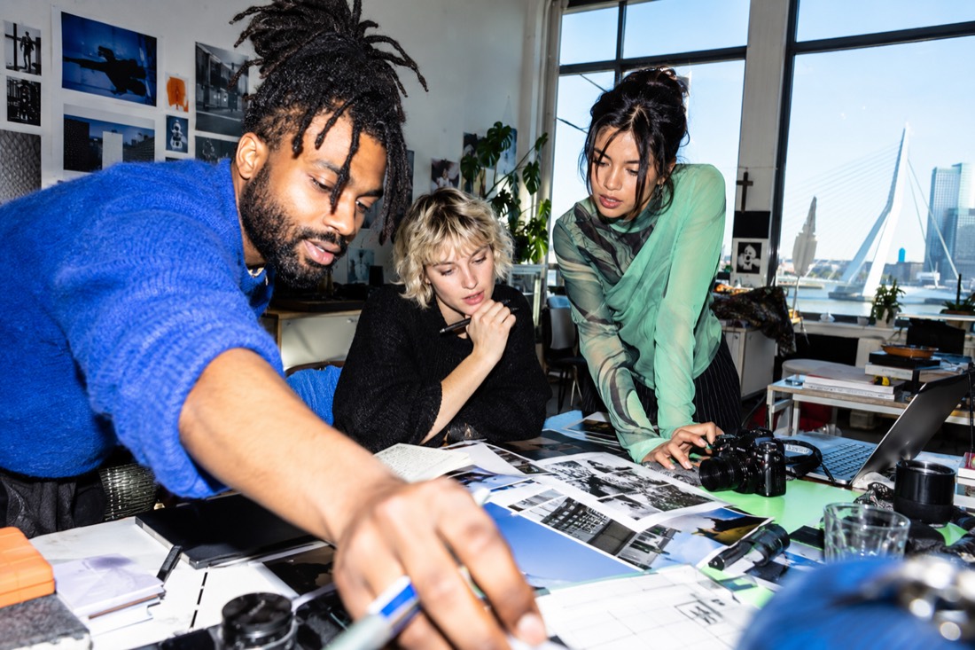 Three people leaning over a table of prints and a camera, the Erasmus bridge through the window behind them
