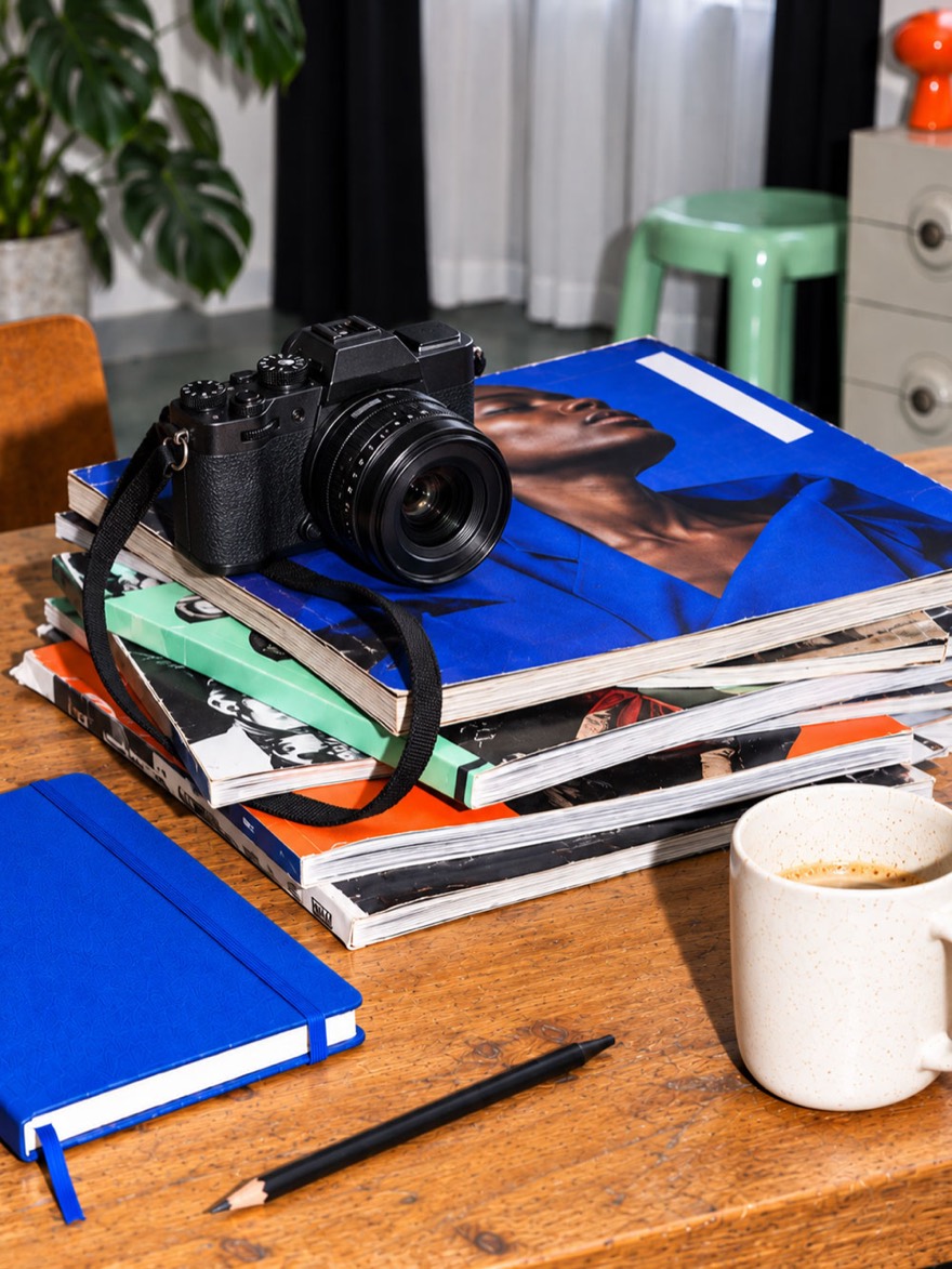 A camera resting on a stack of magazines beside a notebook, a pencil and a cup of coffee